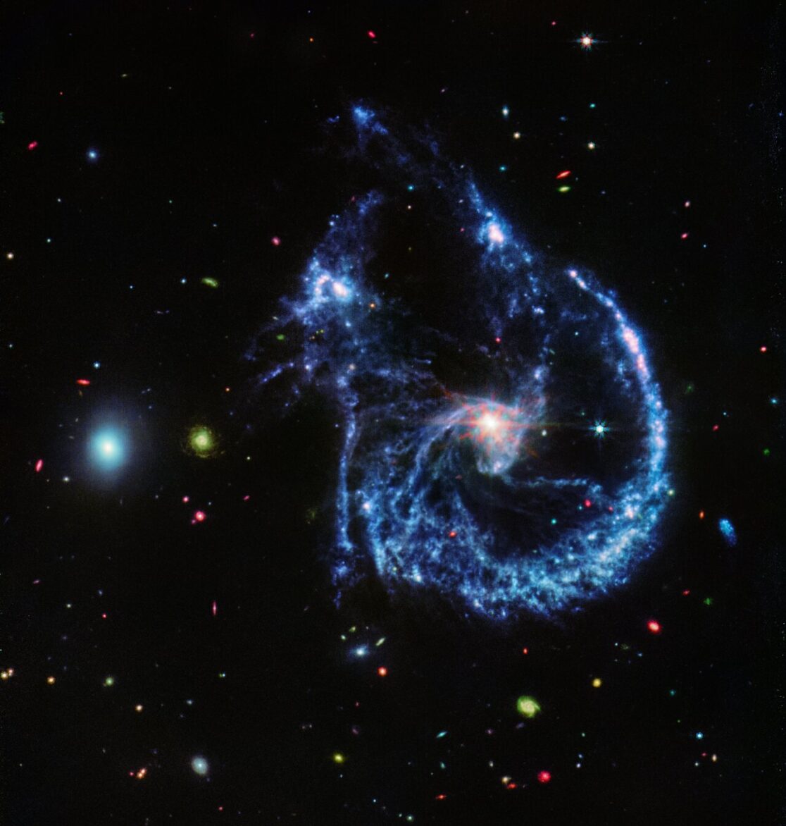 This image of Arp 107, obtained by Webb’s MIRI (Mid-InfraRed Instrument), reveals the supermassive black hole that lies in the centre of the large spiral galaxy to the right, as evidenced by the small, bright central ‘core’. This bright core, where the black hole is pulling much of the dust into lanes, also features Webb’s characteristic diffraction spikes, caused by the light that it emits interacting with the structure of the telescope itself.